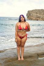 Model wearing Honor The House Breeze Off The Ocean Bikini in Caribbean Cherry colorway with her left arm behind her back standing on a beach shore.